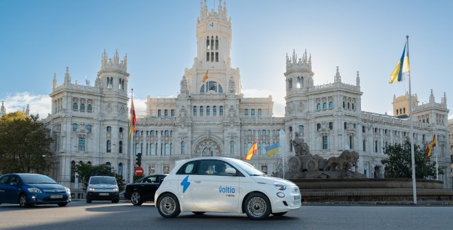 Auto eléctrico compartido marca Voltia circulando frente al majestuoso Palacio de Cibeles en Madrid, con la fuente de Cibeles y banderas visibles en el entorno, bajo un cielo despejado.