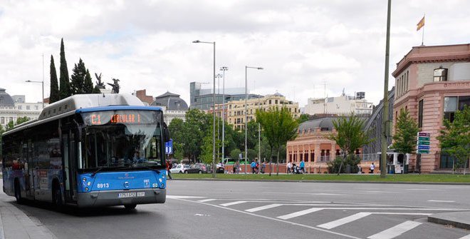 Autobús azul del transporte público de Madrid circulando por una amplia avenida, con árboles, edificios y cielo nublado en el fondo.