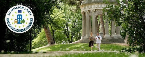 Dos personas caminando en un parque verde y arbolado frente a un monumento clásico con columnas en Madrid. A la izquierda, un sello de la Misión de la UE para ciudades climáticamente neutras y saludables.