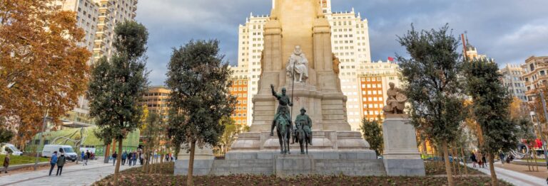 Monumento de Don Quijote y Sancho Panza en la Plaza de España, Madrid, rodeado de árboles y edificios altos en el fondo bajo un cielo parcialmente nublado.